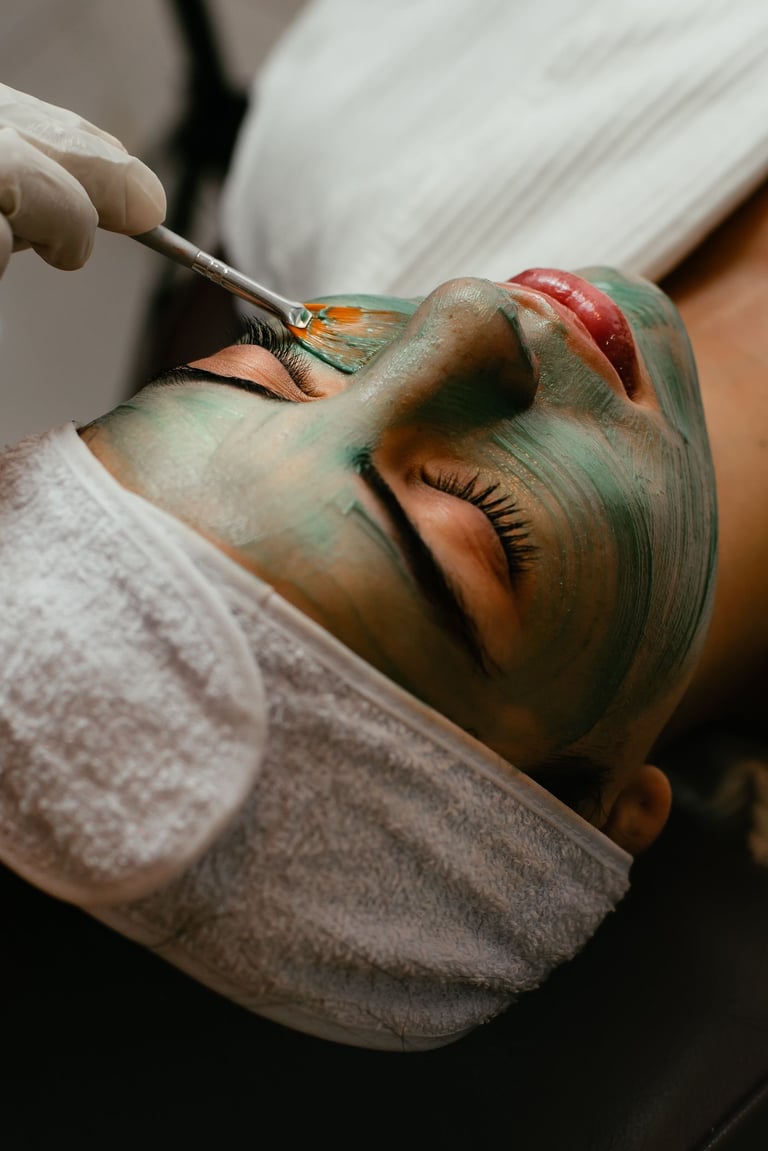 Esthetician applying green facial mask with brush to clients face, with red lip treatment visible